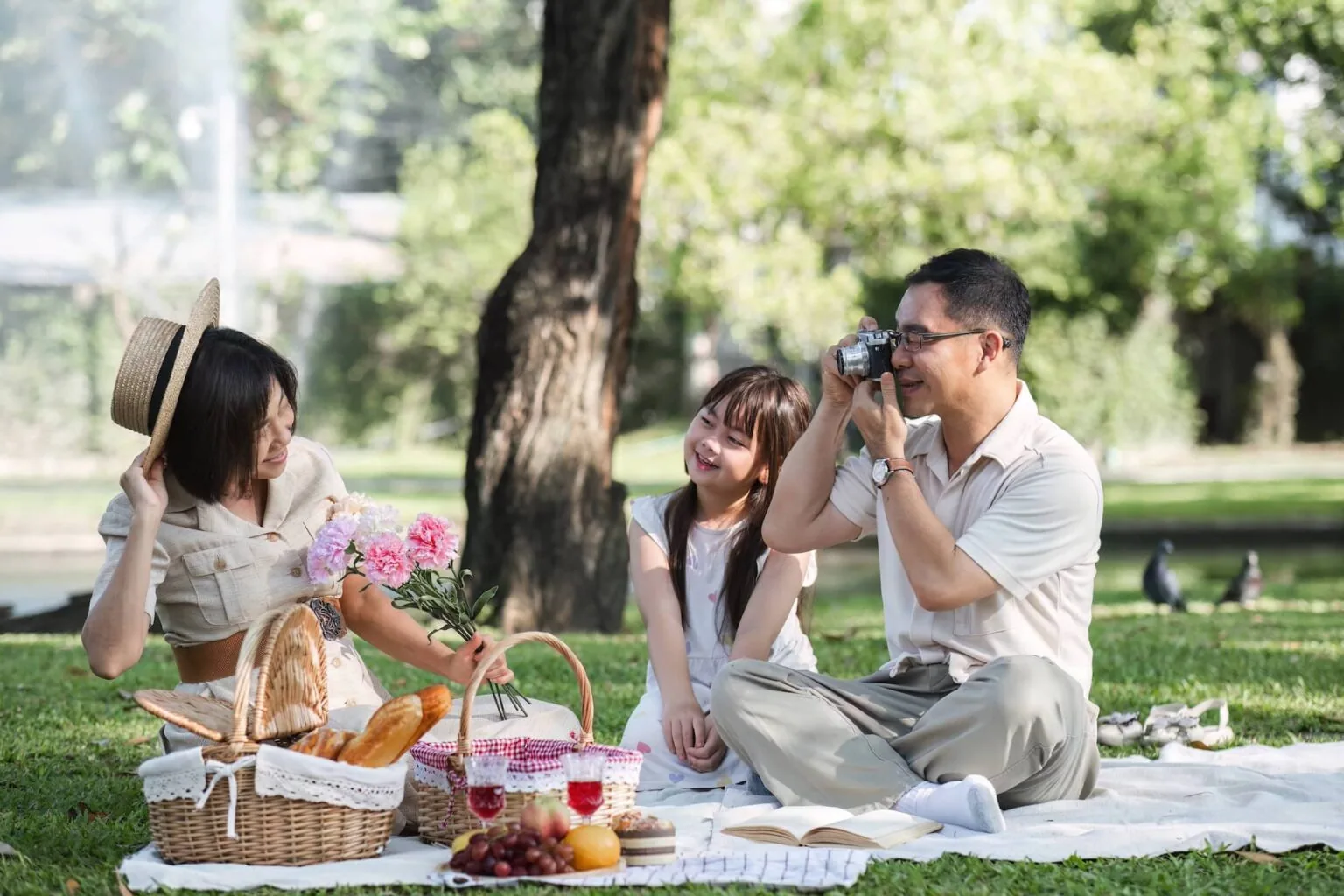 Family of three on picnic blanket in park, father photographing with camera while mother and daughter smile