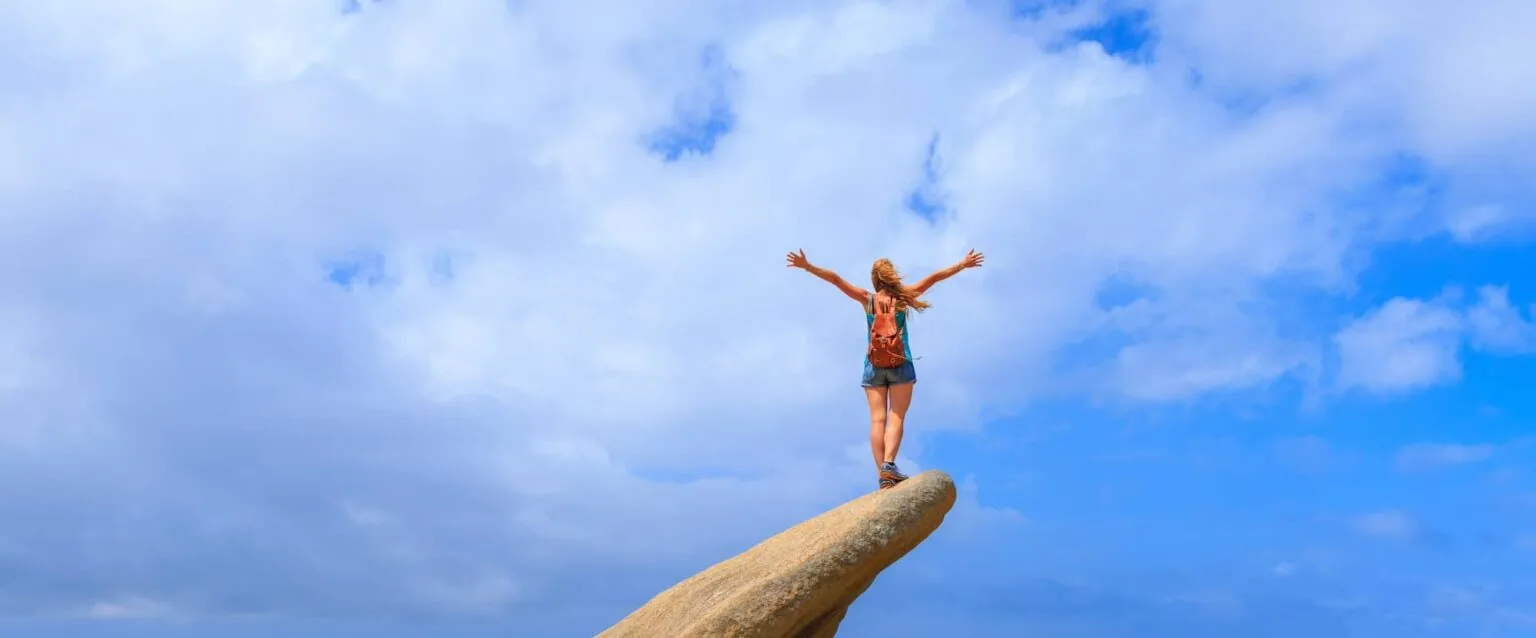 Person standing on a wooden pole with arms raised against a blue sky with white clouds